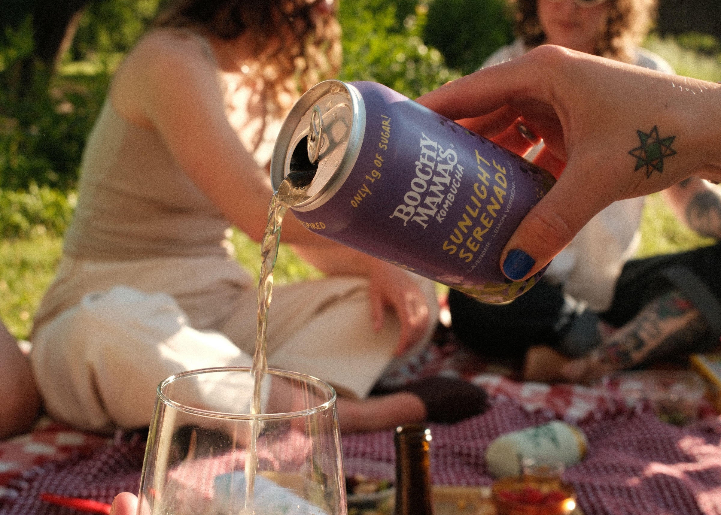 A purple can of kombucha is poured into a glass above a picnic blanket with two friends sitting in a sunny backdrop. 